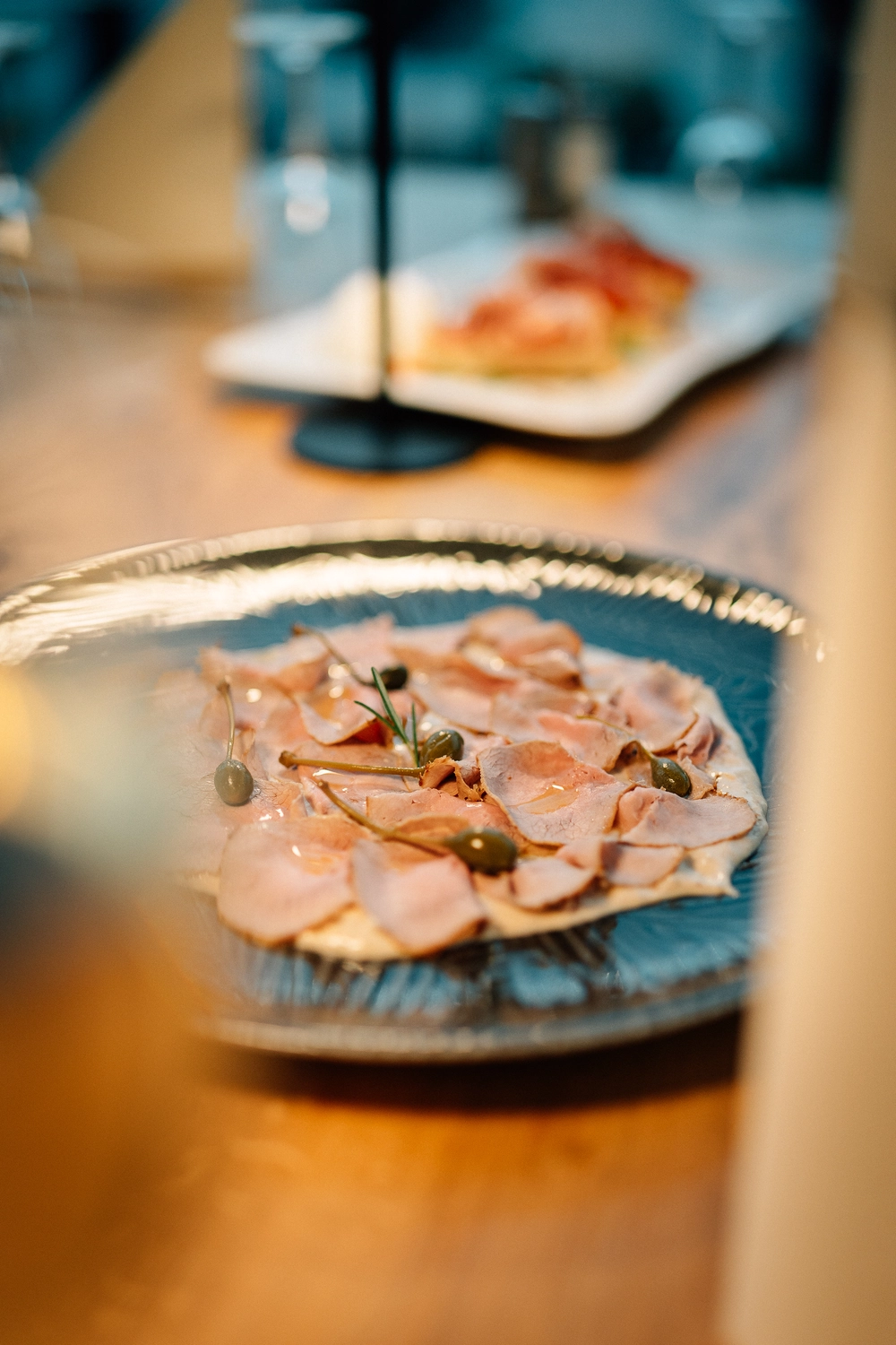 Sinn Kitchen – Restaurant – Ein Koch beträufelt dünn geschnittenes Fleisch, wahrscheinlich Bestandteil eines italienischen Gerichts wie Vitello Tonnato oder Prosciutto, mit einer Flüssigkeit, vermutlich Olivenöl oder einem Dressing.